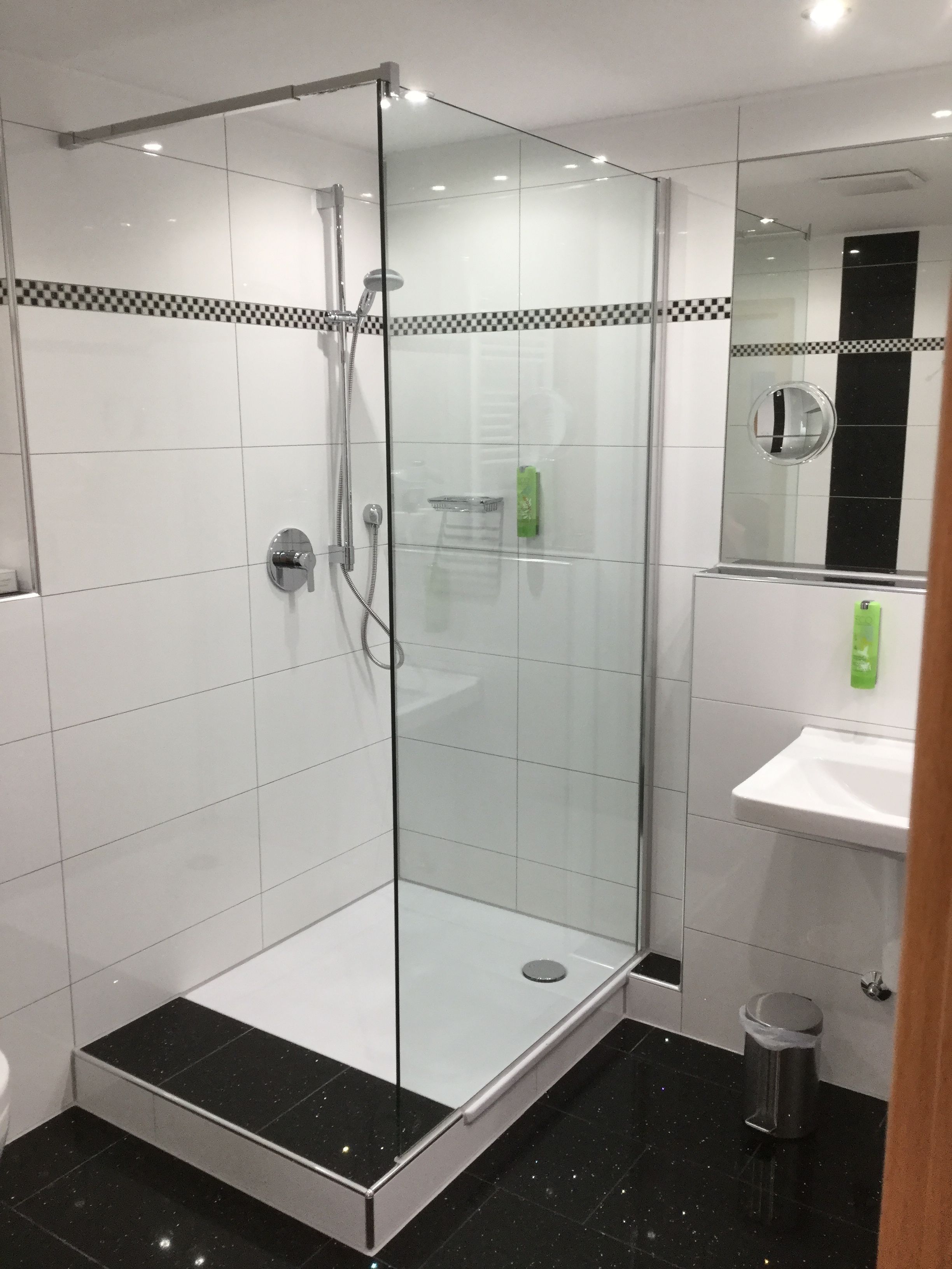 A modern bathroom with a glass-enclosed shower, white tiled walls, a black-tiled floor, and a white sink with a mirror above it.