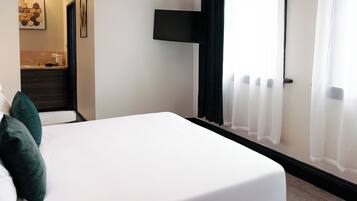 Habitación de hotel moderna con una cama bien hecha, una pequeña zona de estar, aire acondicionado de pared y vistas al exterior a través de cortinas traslúcidas.