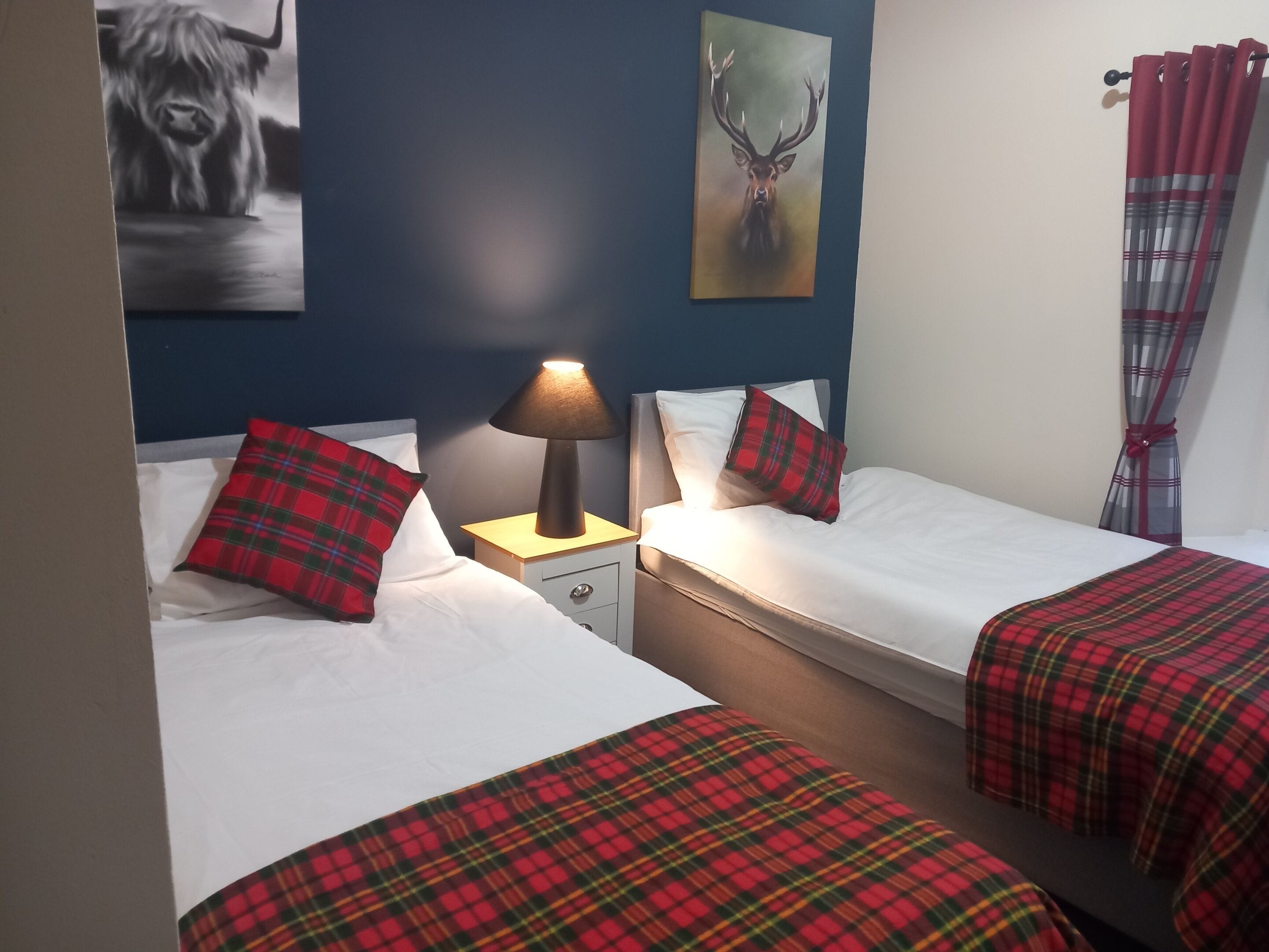 A bedroom with two beds, each with red and green plaid bedding, a bedside lamp, and a painting of a deer on the wall.