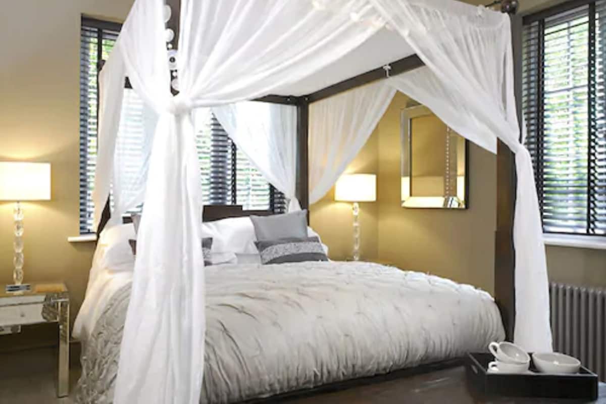 A four-poster canopy bed with a white bedspread and pillows, bedside lamps, a nightstand, and a tray with cups and a saucer.