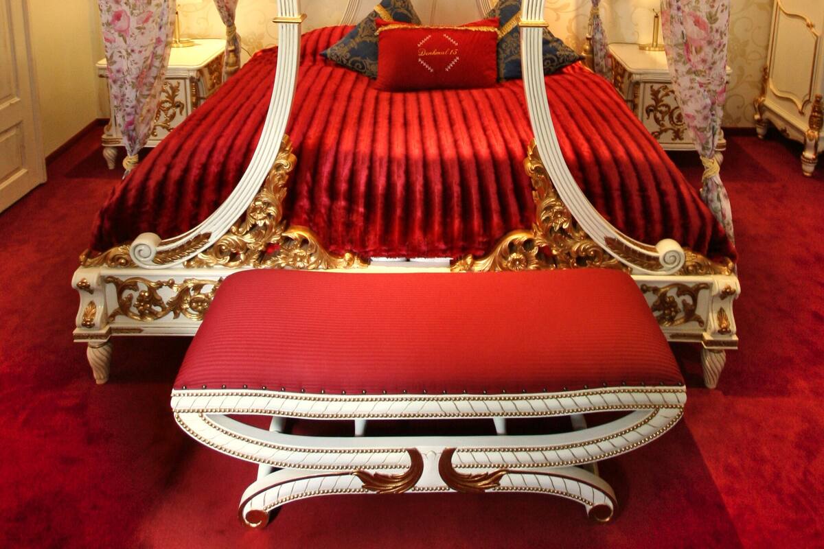 Ein Himmelsbett mit roter Polsterung und goldenen Akzenten, eine Bank am Fußende sowie blumige Vorhänge.