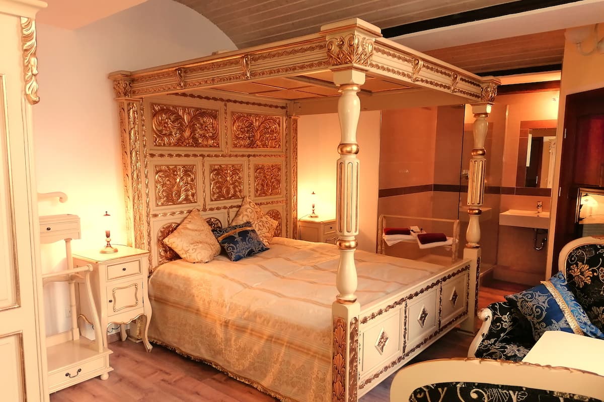 Ein Bett mit Baldachin, verziertem Kopfteil und Fußteil, ein Nachttisch mit Lampe sowie ein Sessel mit Blumenmuster.