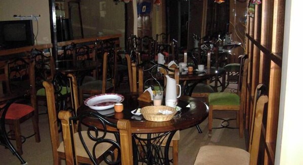 Un comedor con varias mesas y sillas de madera, algunas con cojines. Hay platos, cuencos y una jarra en las mesas.