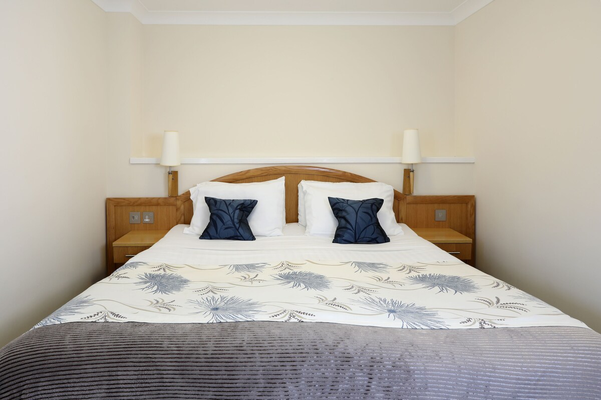 Una cama bien tendida con colcha estampada, dos almohadas a juego y un cabecero de madera. Hay dos mesitas de noche con lámparas, y las paredes están pintadas de un tono claro.