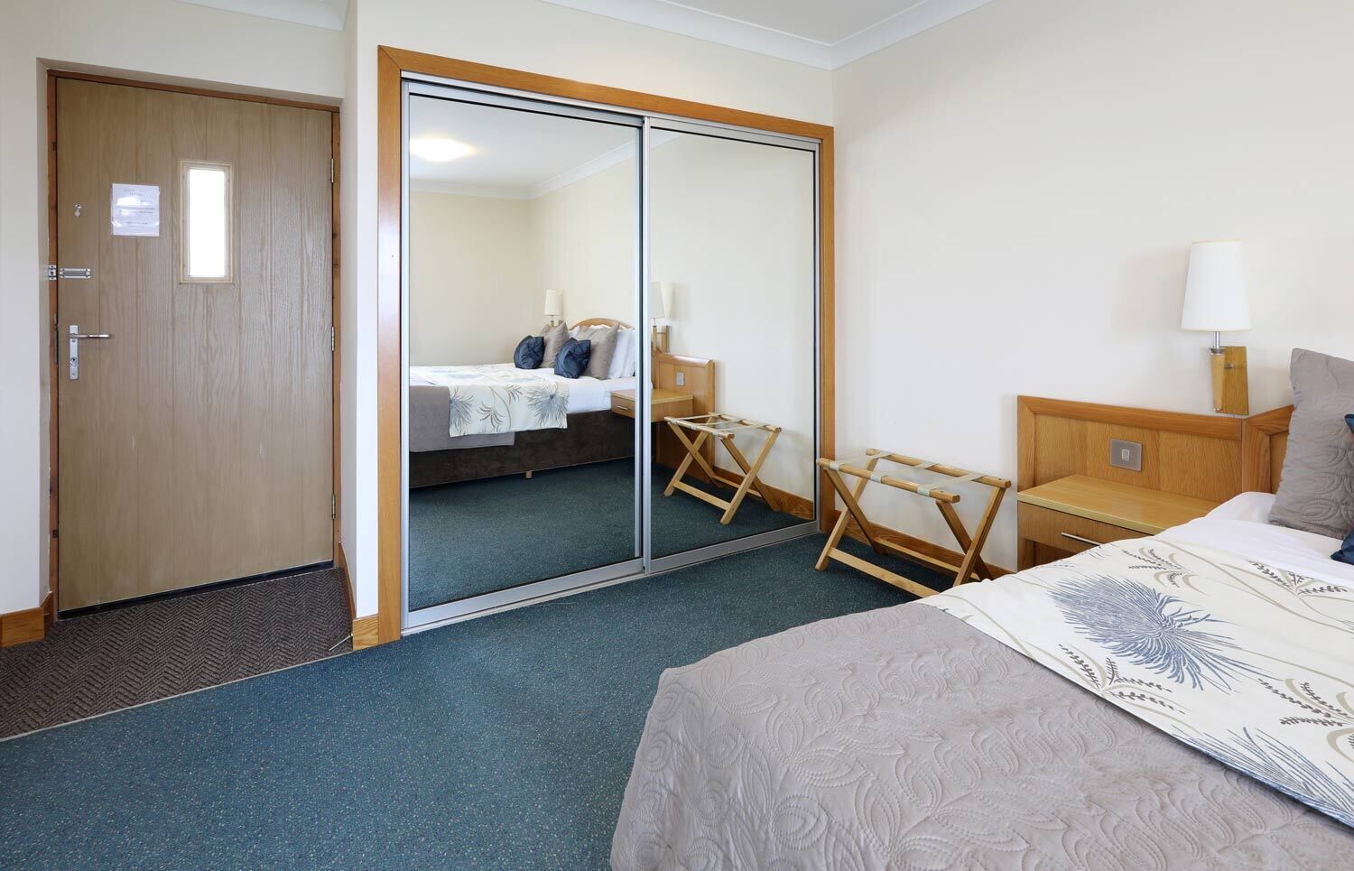 Una habitación de hotel con cama, una mesita de noche de madera, una silla, un espejo y una puerta.