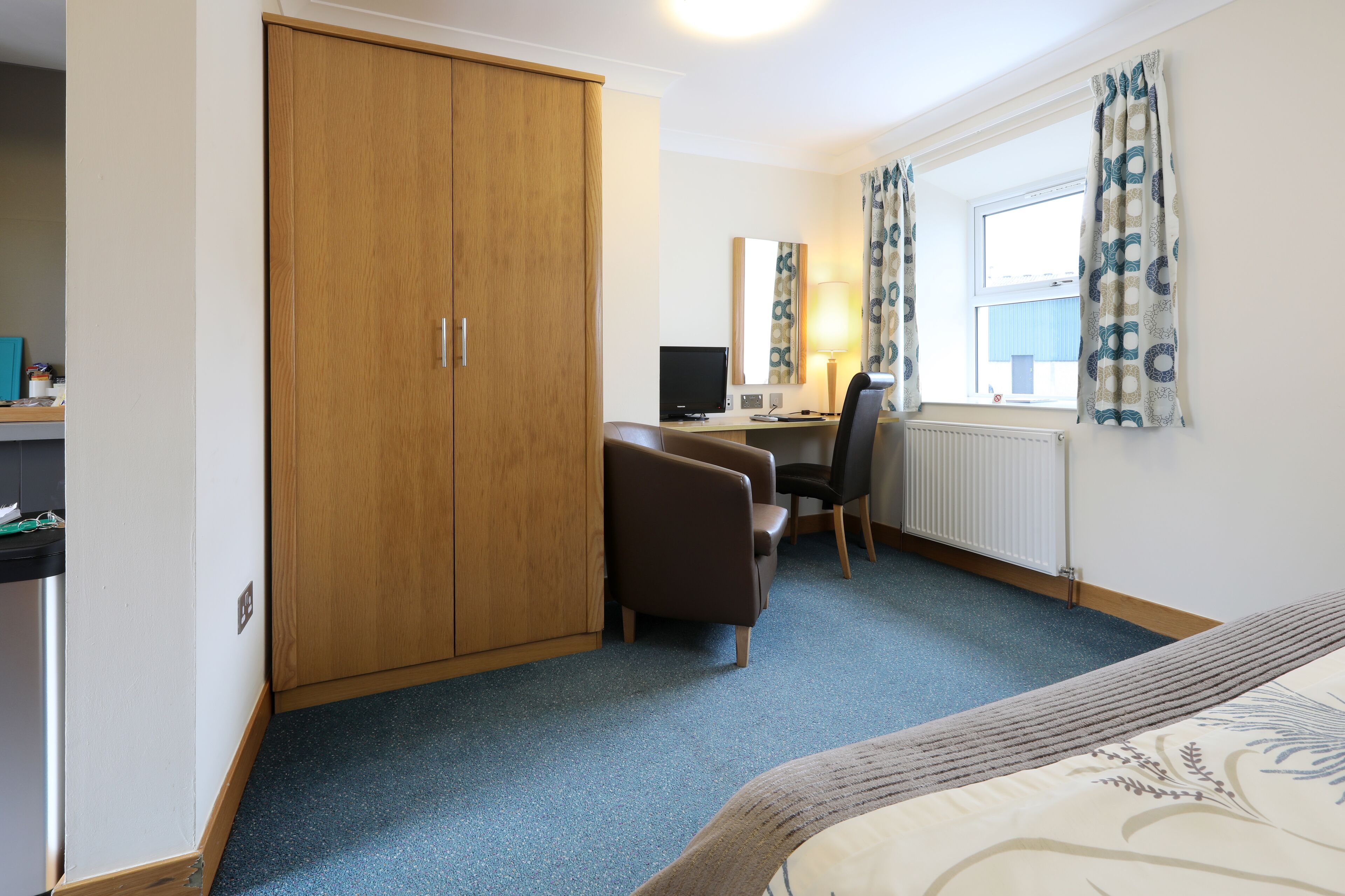 A hotel room with a wardrobe, a desk with a computer, a chair, a bed, and a window with curtains.