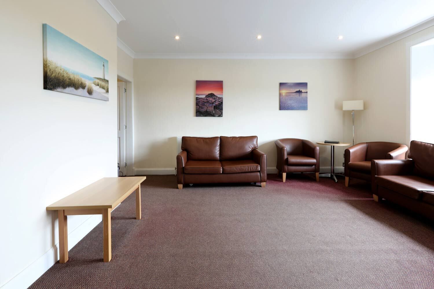 A spacious living room with a brown leather sofa, a wooden coffee table, and two armchairs. There are three paintings on the wall and a floor lamp.