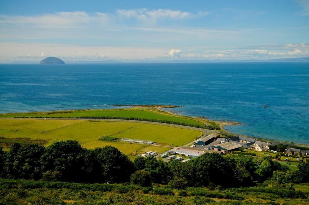Un paysage côtier avec une étendue d’eau importante, une petite île au loin, et un mélange de champs verdoyants et de bâtiments.