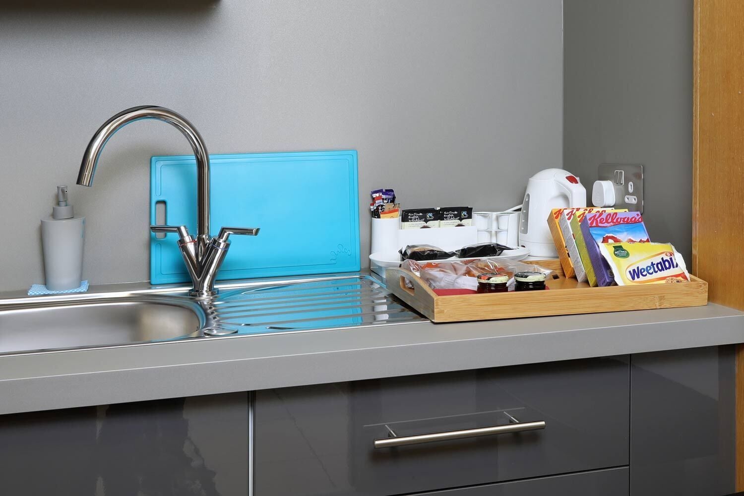 A modern kitchen with a stainless steel sink, a blue soap dispenser, a coffee maker, and a tray with snacks.