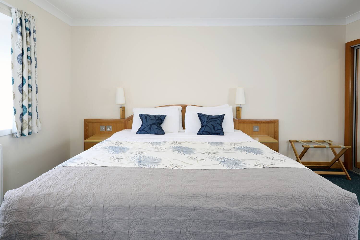 A hotel room with a large bed, two bedside tables with lamps, a wooden headboard, and a window with curtains.