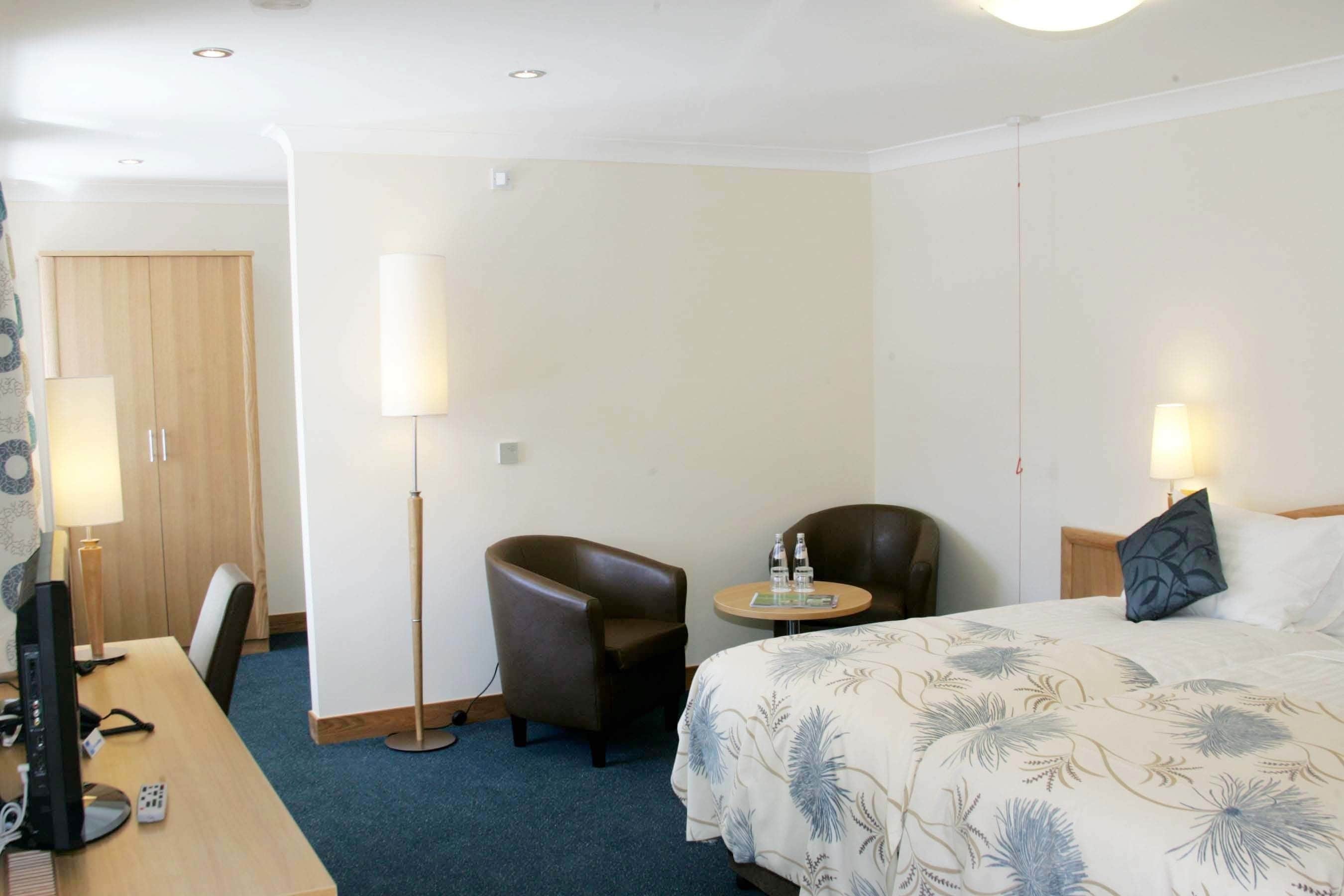 Une chambre d’hôtel comprenant un lit, deux fauteuils, un bureau avec un ordinateur et deux lampes de chevet.