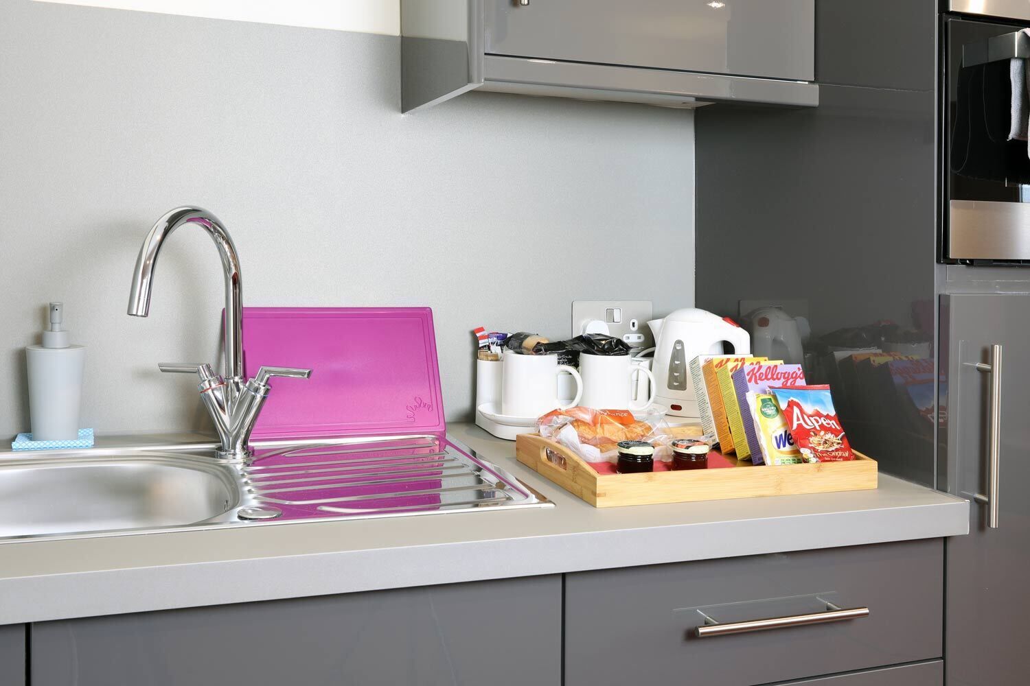 A modern kitchen with a stainless steel sink, a pink drying rack, a coffee maker, and a breakfast tray with cereal and spreads.