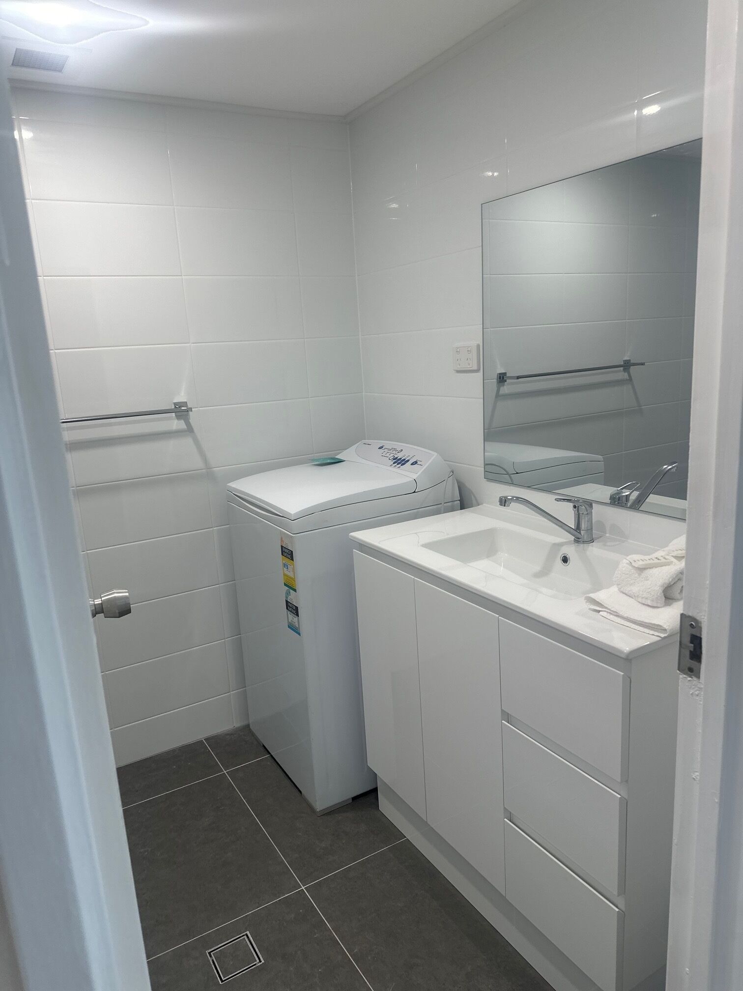 A small, modern bathroom with white tiles, a built-in washing machine, a sink with a mirror, and a towel rack.