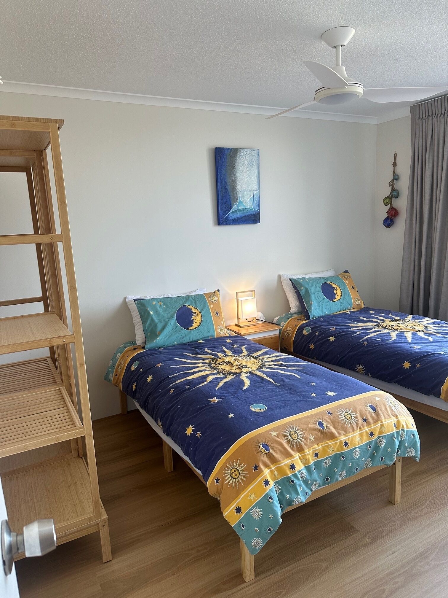 Une chambre avec deux lits, une étagère en bois, un ventilateur de plafond, un tableau mural et un rideau.