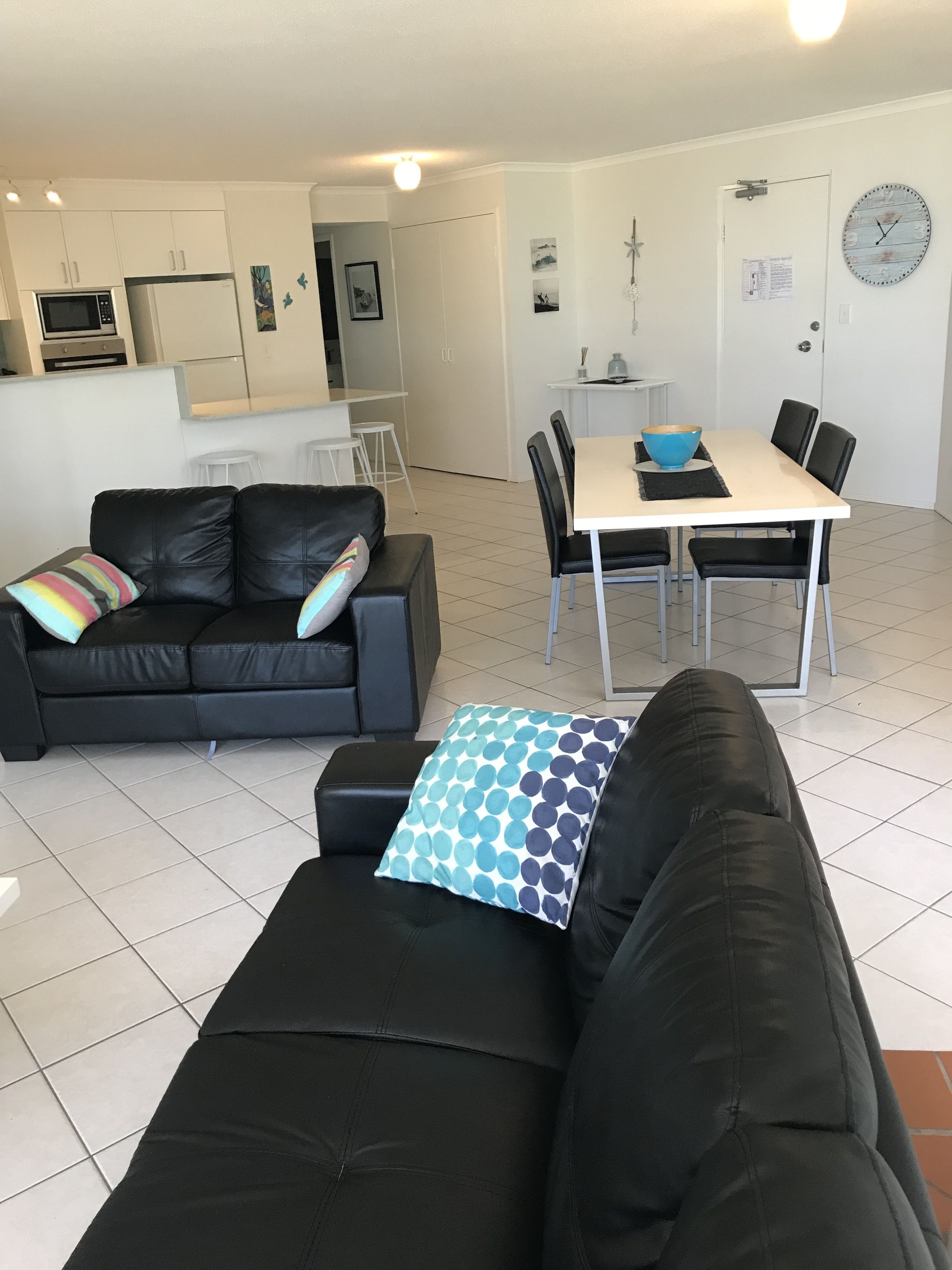 A modern living room with a black leather sofa, a dining table with chairs, and a kitchen area in the background.