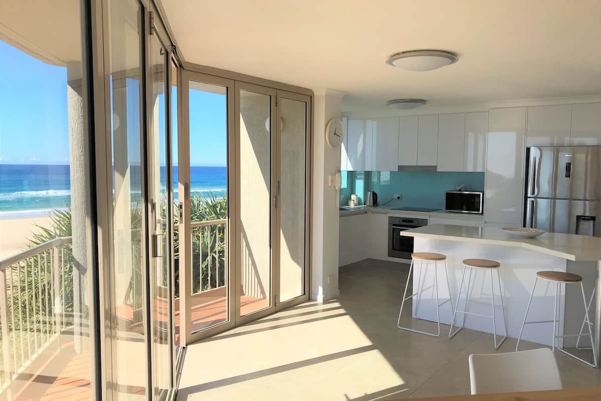 A modern kitchen with a view of the ocean, a balcony with a glass door, and a dining area with stools.