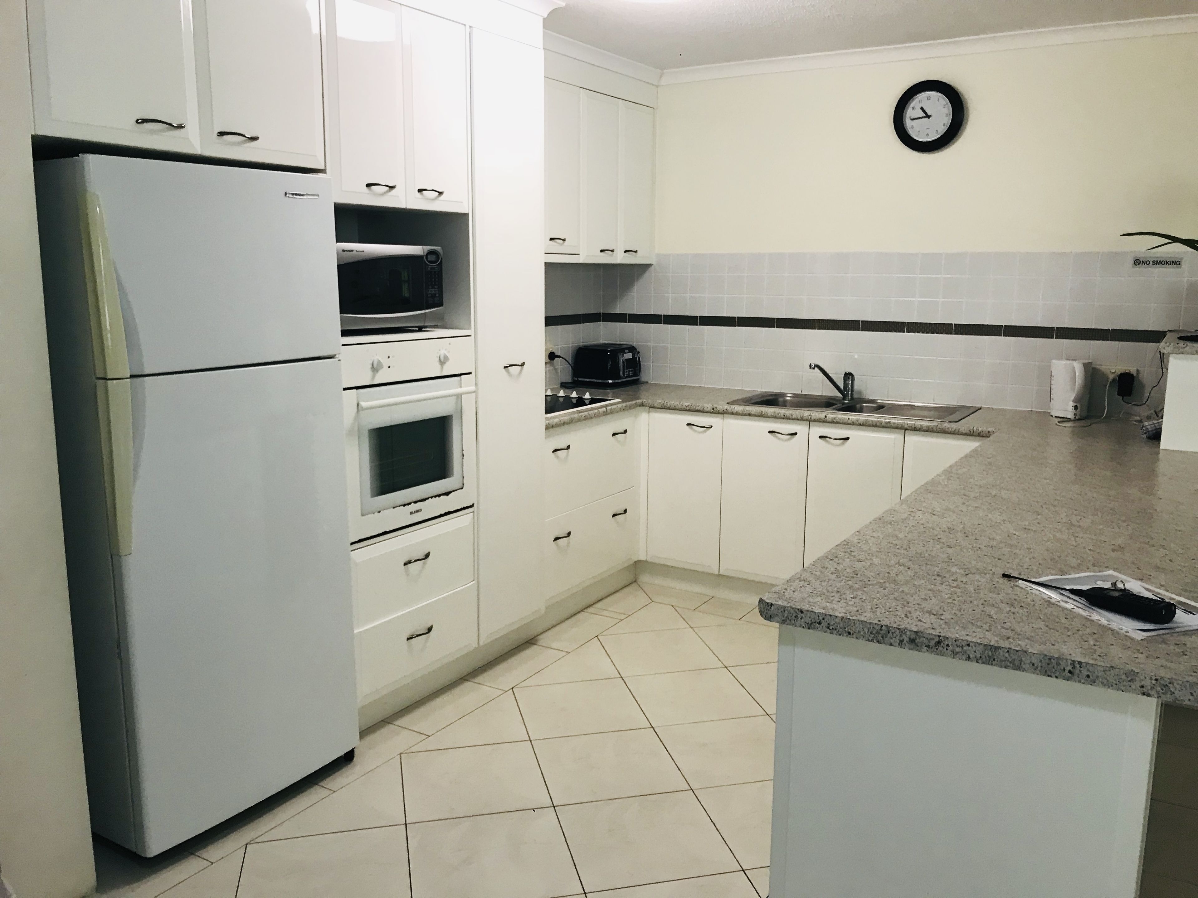 A kitchen with white cabinets, a refrigerator, a microwave, an oven, a sink, and a clock on the wall.