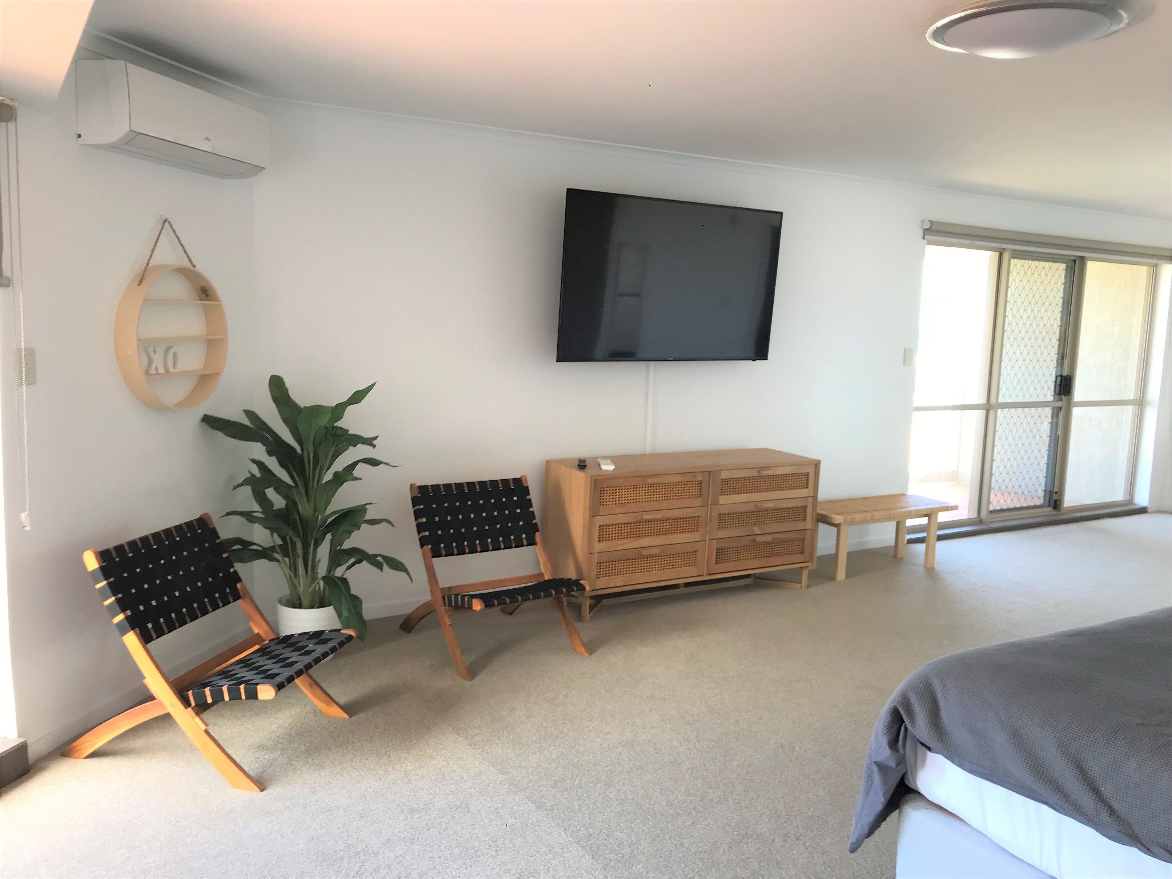 A bedroom with a bed, a TV mounted on the wall, a wooden dresser, two chairs, a potted plant, and a sliding glass door.