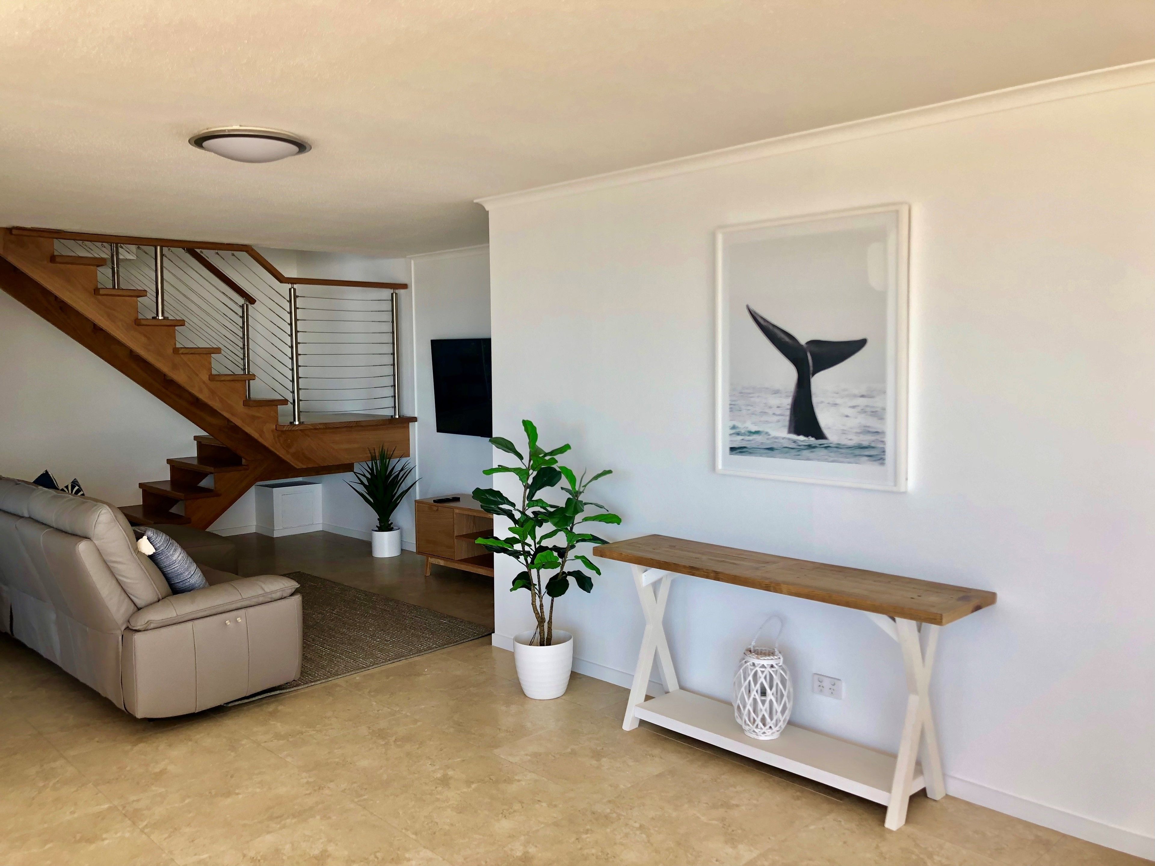 A modern living room with a beige sofa, a wooden staircase, a white console table, and a whale tail artwork on the wall.