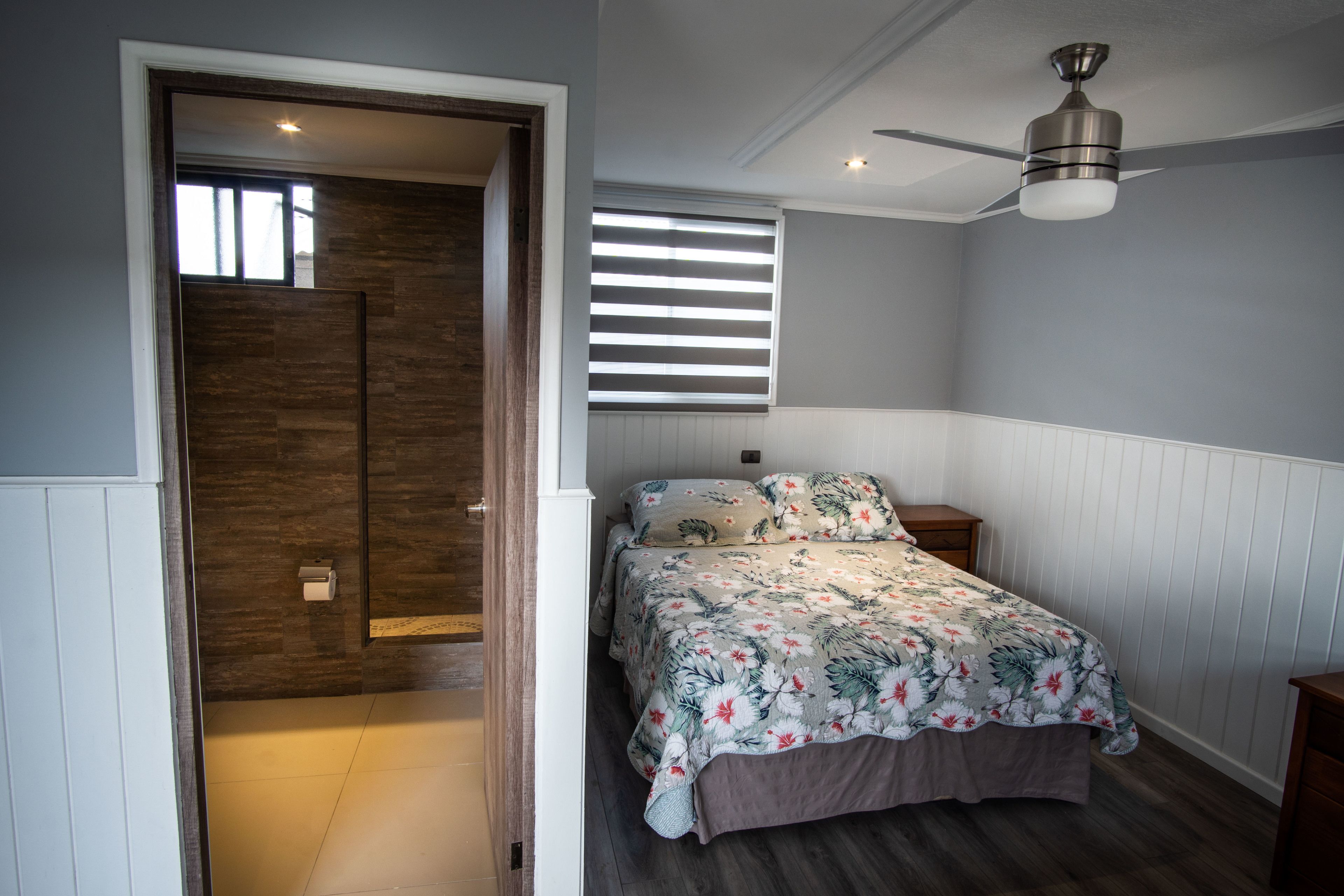 Un dormitorio con cama, ventilador de techo, ventana con persianas y puerta que da a otra habitación.