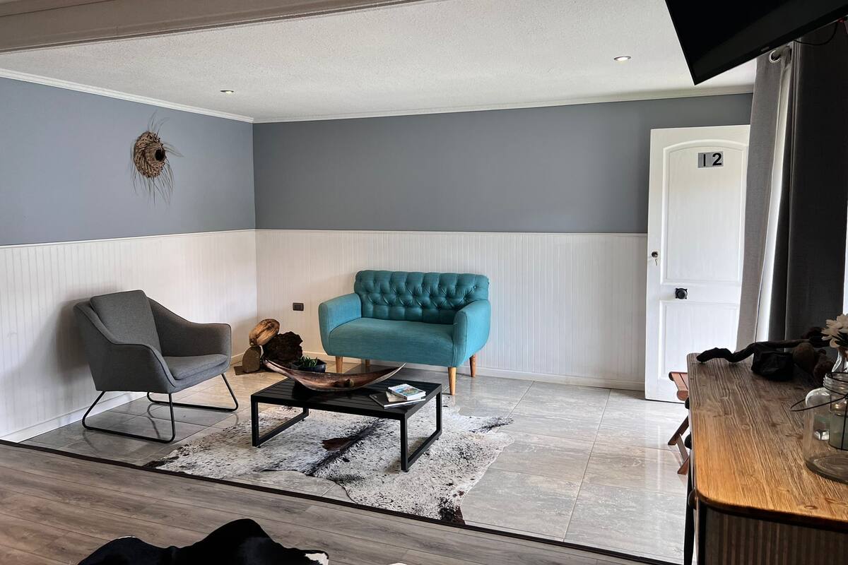 A modern living room with a grey armchair, a teal sofa, and a coffee table. A wooden door with the number 12 is visible.