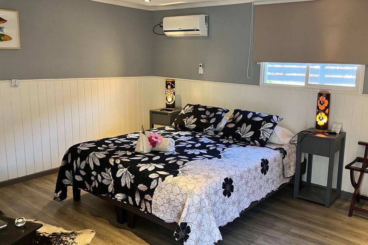 A bedroom with a bed, bedside tables, an air conditioner, a window, and a nightstand with a lamp.