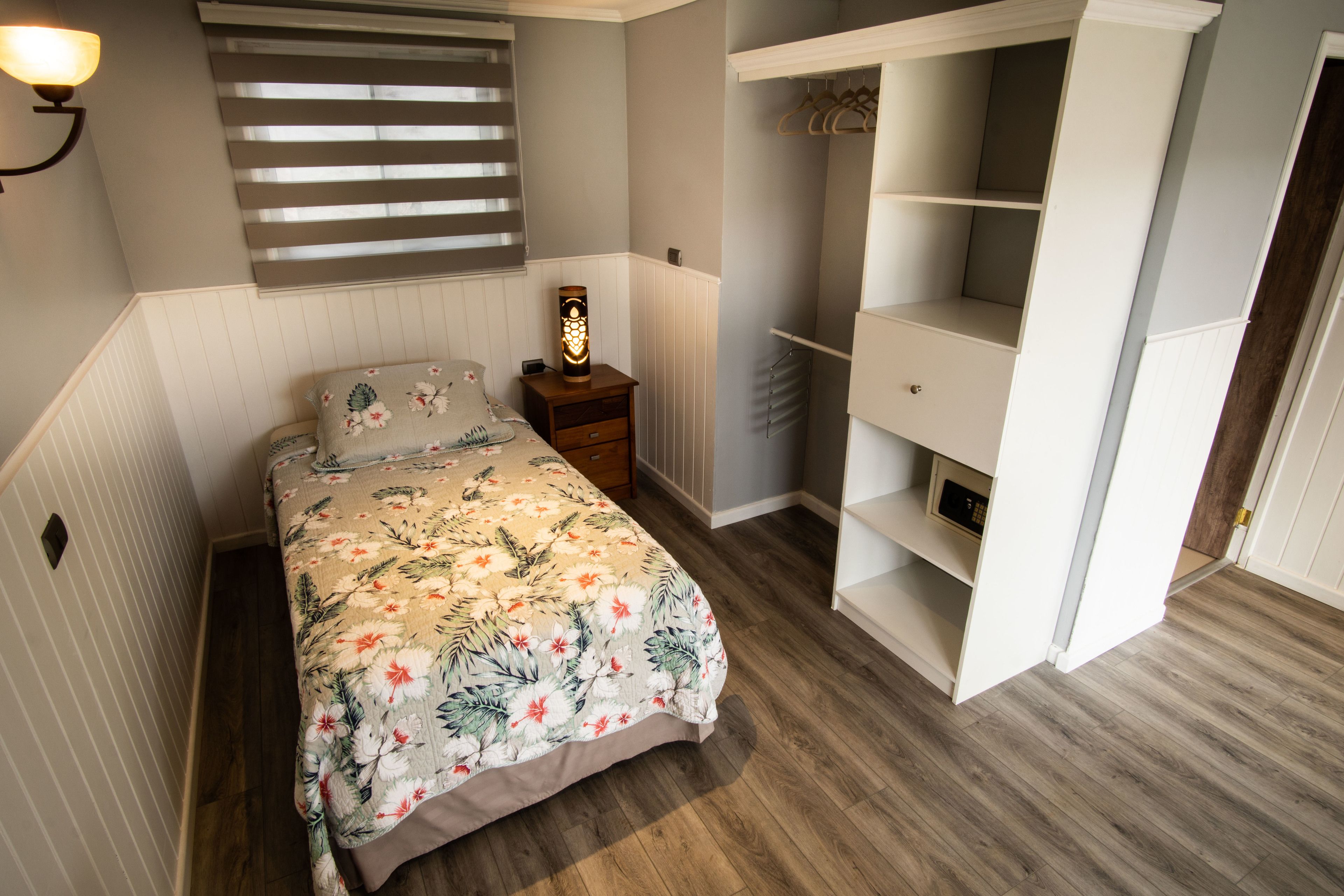 Un dormitorio con cama, mesita de noche, luz empotrada en la pared, ventana con persianas y armario con estantes.