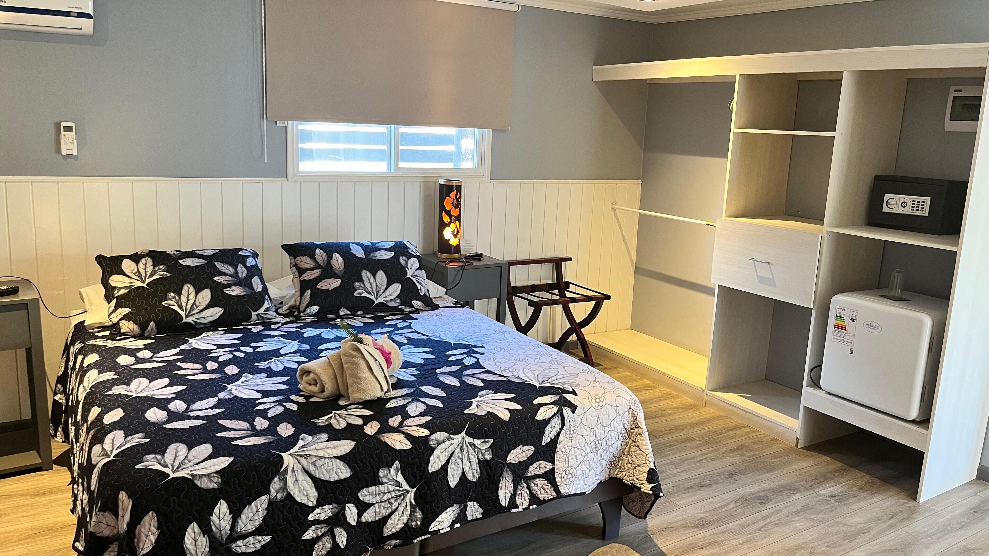 A bedroom with a bed, a bedside table, a desk, a chair, a wardrobe, and a small refrigerator.