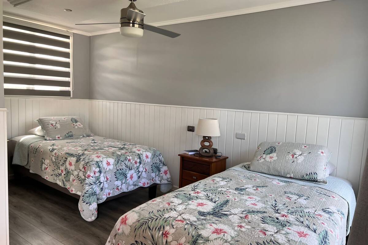A bedroom with two beds, a ceiling fan, a nightstand with a lamp, and a window with blinds.