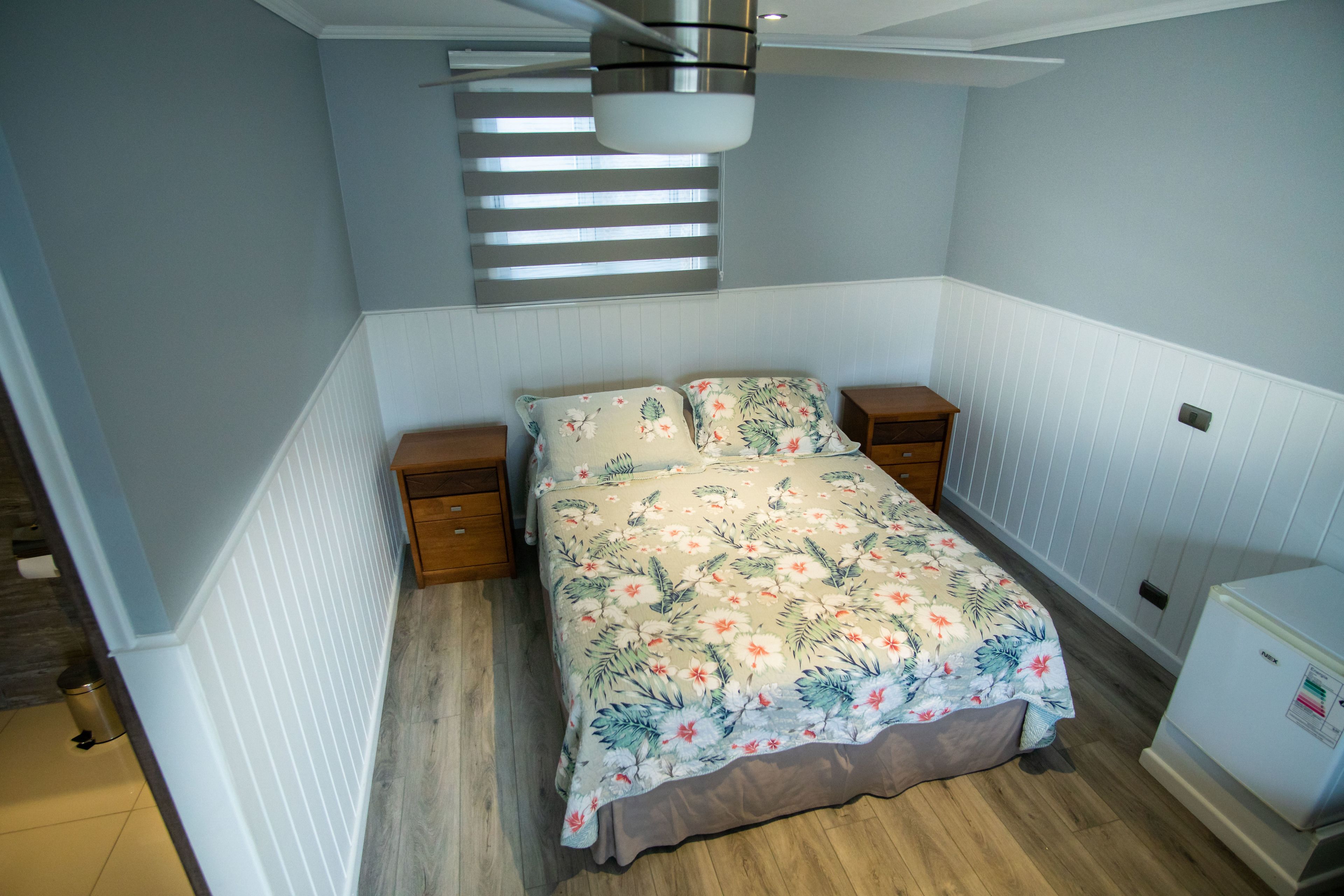Een slaapkamer met een bed, nachtkastjes, een plafondventilator, een raam met jaloezieën en een klein wit apparaat.