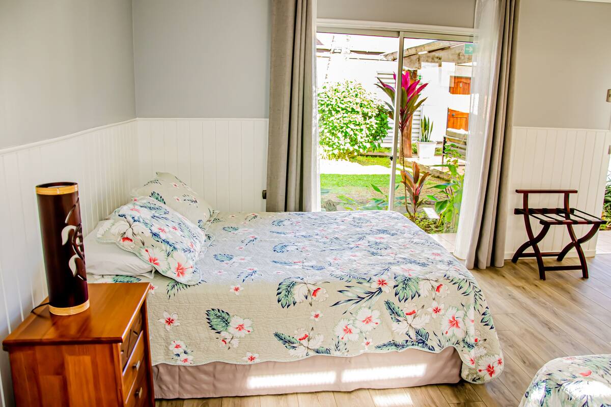 A bedroom with a bed, a wooden nightstand, a chair, and a view of the garden through the window.