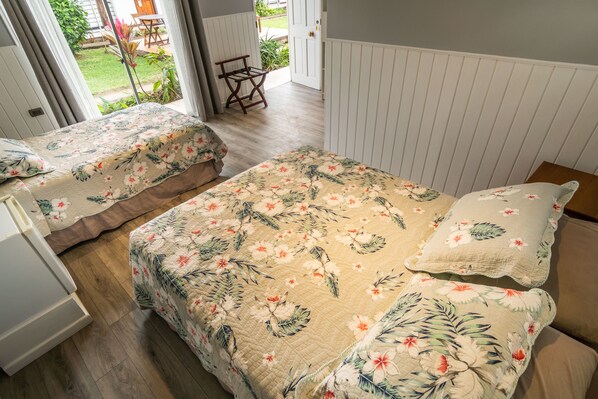 A bedroom with two beds, wooden flooring, and a view of the garden through an open door.