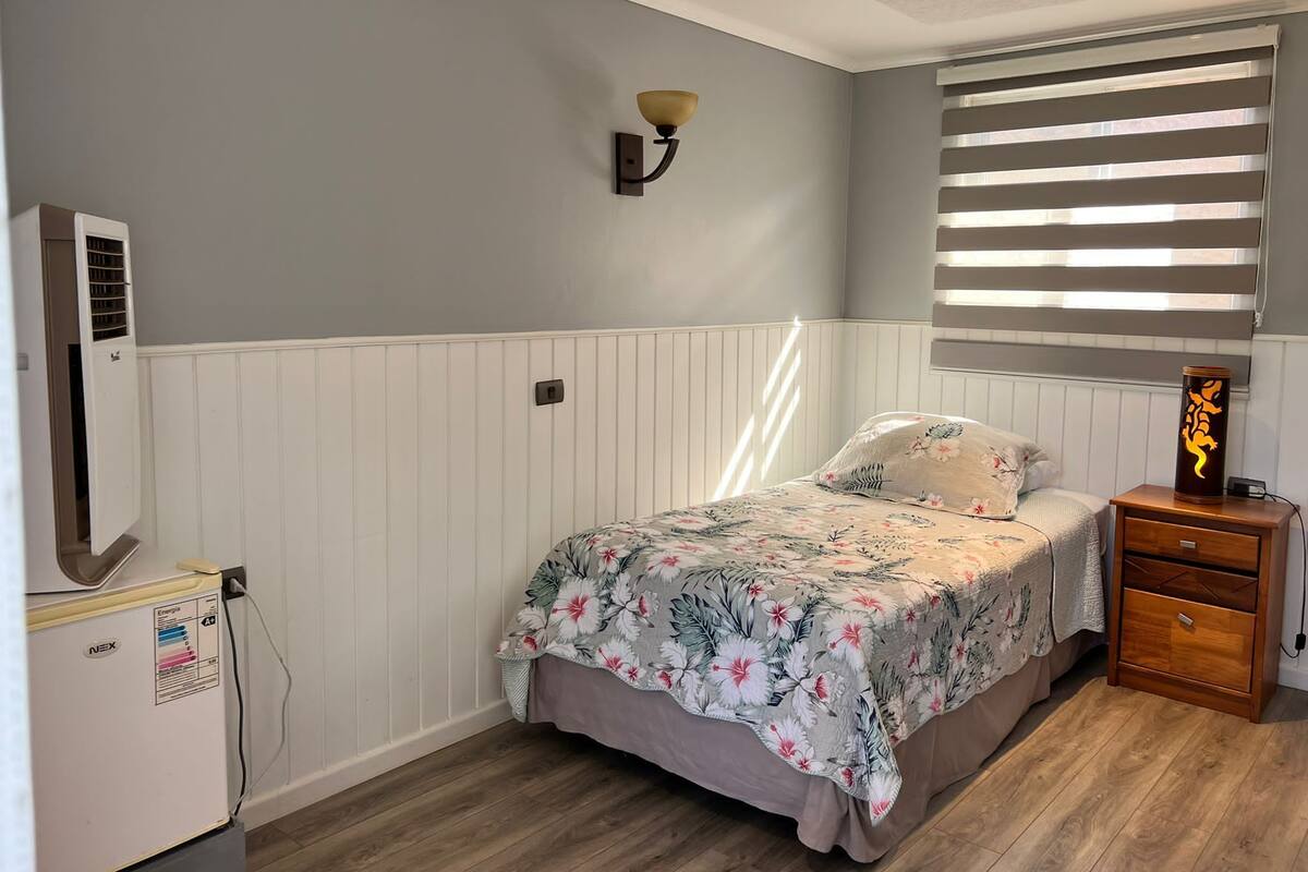 A bedroom with a bed, a nightstand, a wall-mounted lamp, a window with blinds, and a wall-mounted air conditioner.
