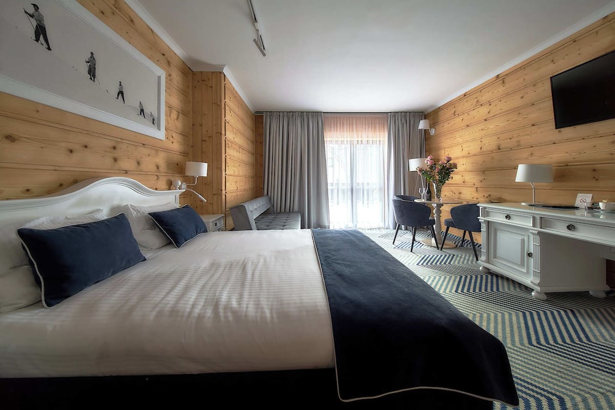 A hotel room with a large bed, a desk, two chairs, a flat-screen TV, and a window with curtains.