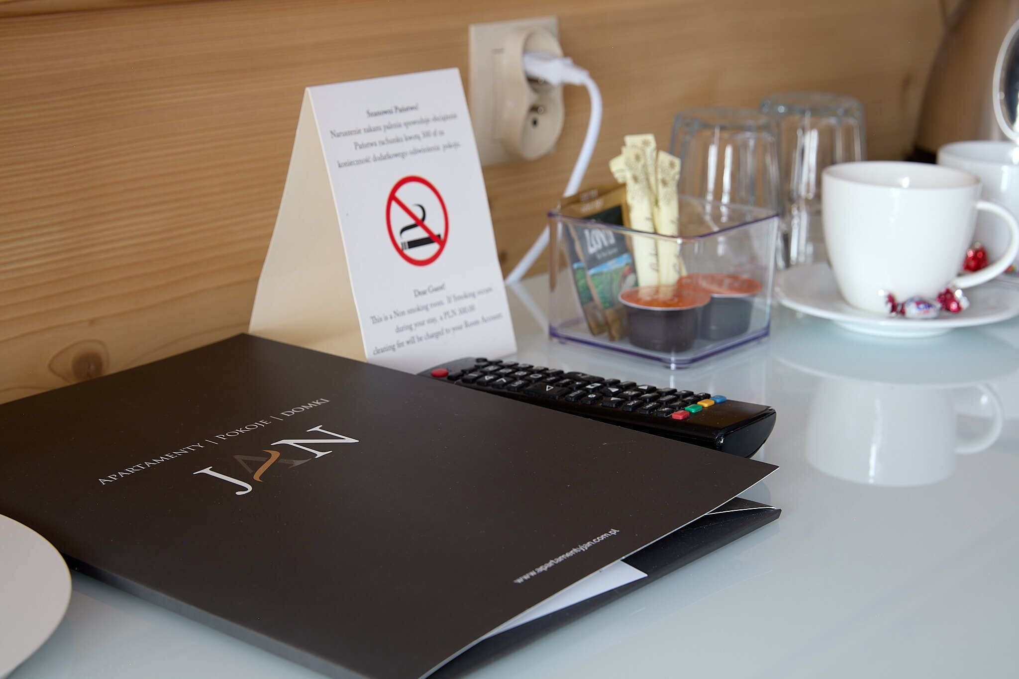 Ein Hotelzimmer mit einem schwarzen Buch mit dem Titel "JAN" auf einem Tisch, einem Schild mit einem Rauchverbotssymbol und einem Glasbehälter mit einer Fernbedienung.