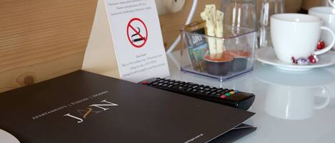 A hotel room with a black book titled "JAN" on a table, a sign with a no-smoking symbol, and a glass container with a remote control.