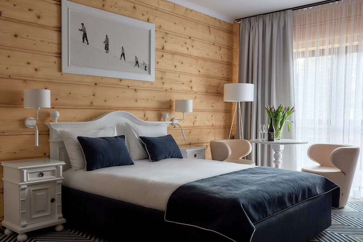 A hotel room with a wooden panel wall, a large bed with dark blue bedding, two bedside tables with lamps, a small round table with a vase of flowers, and a chair.