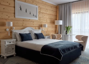 A hotel room with a wooden paneled wall, a large bed with navy bedding, two bedside tables with lamps, a small round table with a vase of flowers, and a chair. - Willa Jan (Zakopane)