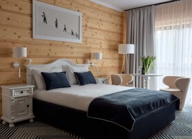 A hotel room with a wooden panel wall, a large bed with dark blue bedding, two bedside tables with lamps, a small round table with a vase of flowers, and a chair.