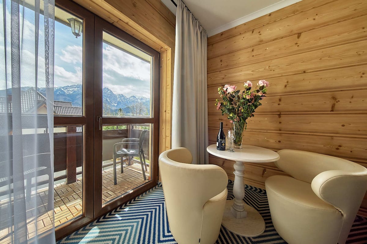 A room with a wooden interior, a glass door leading to an outdoor area with a table and chairs, and a vase with flowers on a small table.