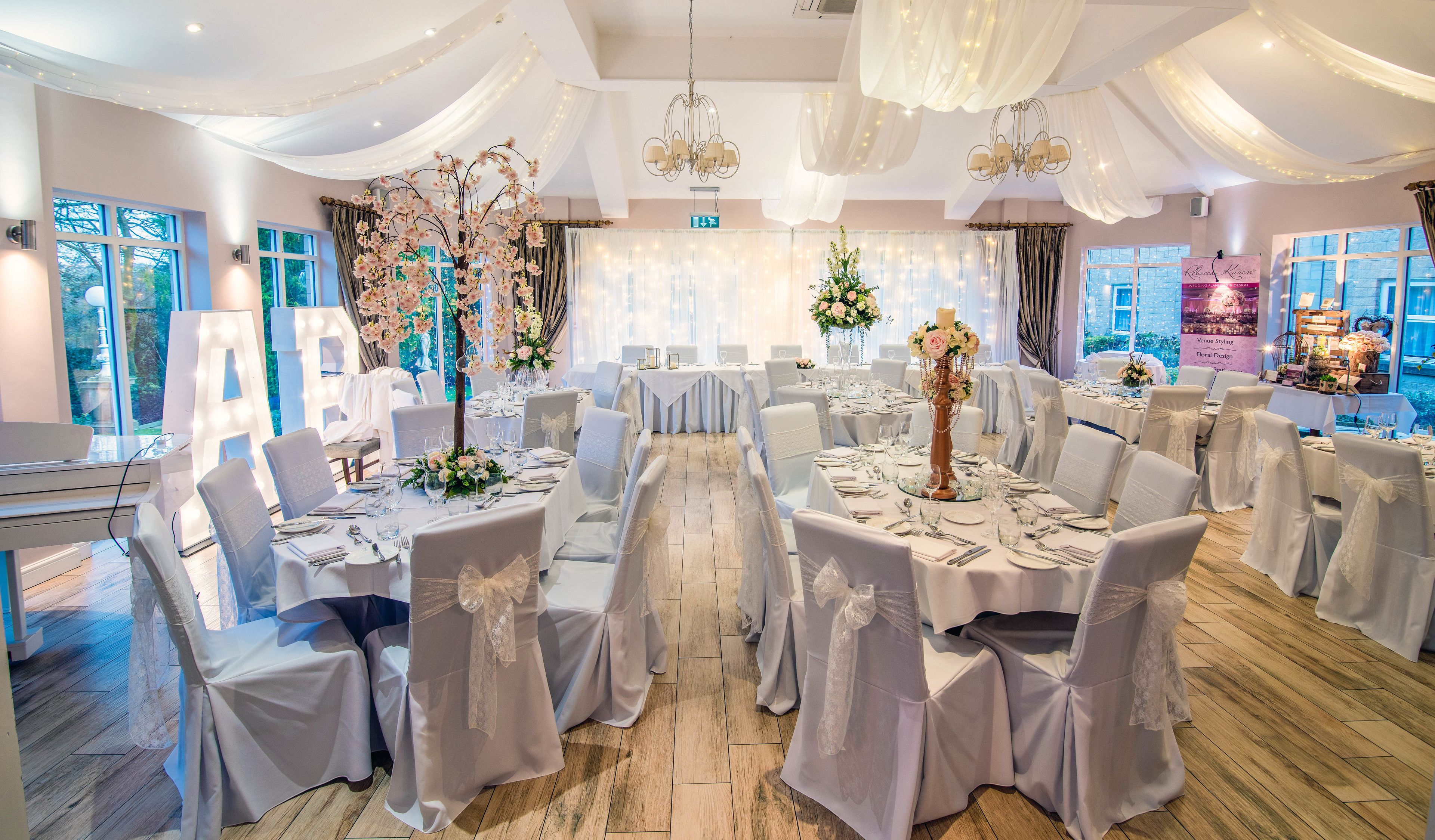 Une salle de banquet où les tables sont dressées pour un événement formel, avec des nappes blanches, des centres de table floraux et des lustres.