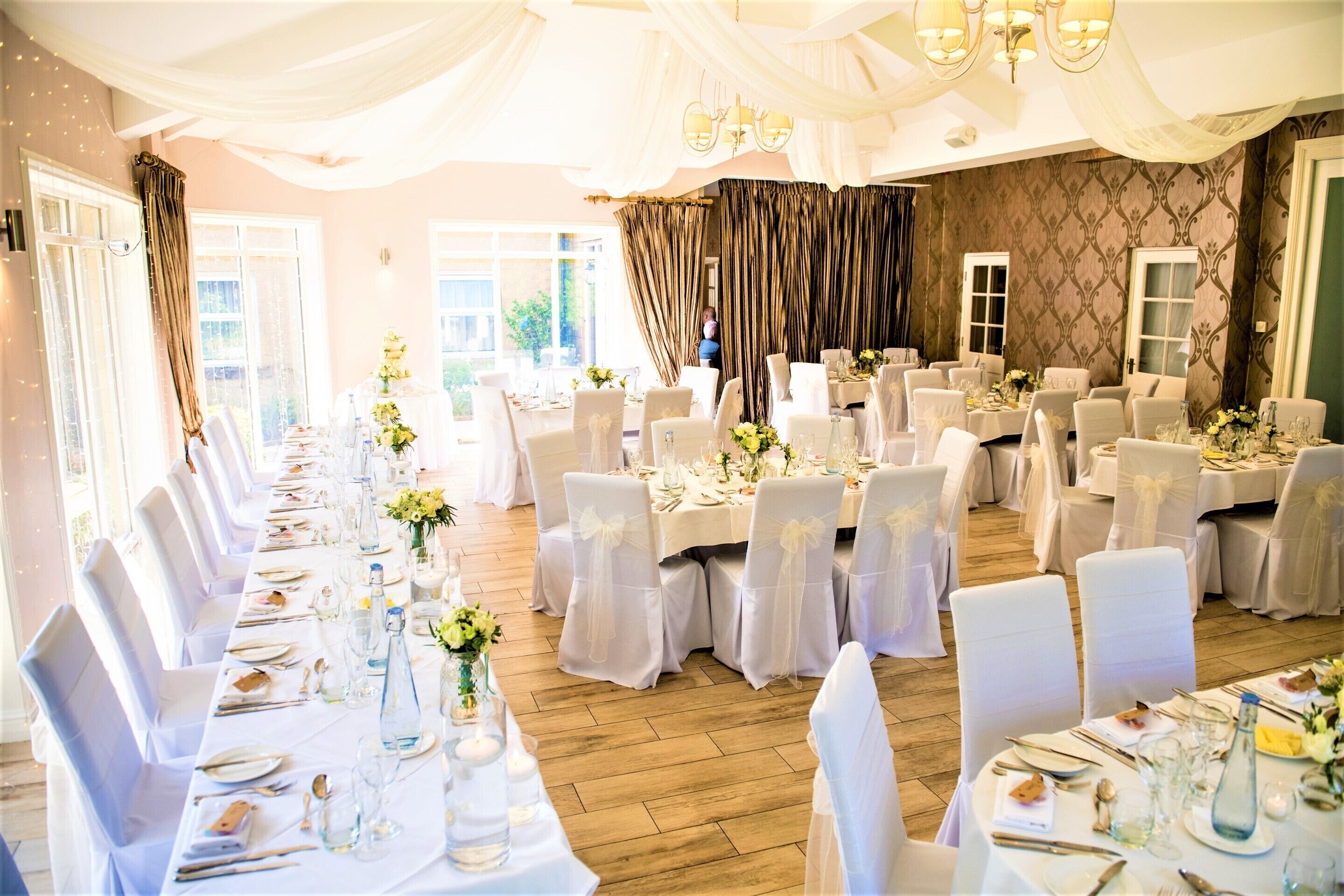 A banquet hall with long tables set for a formal event, featuring white tablecloths, chairs with white covers, and floral centerpieces.