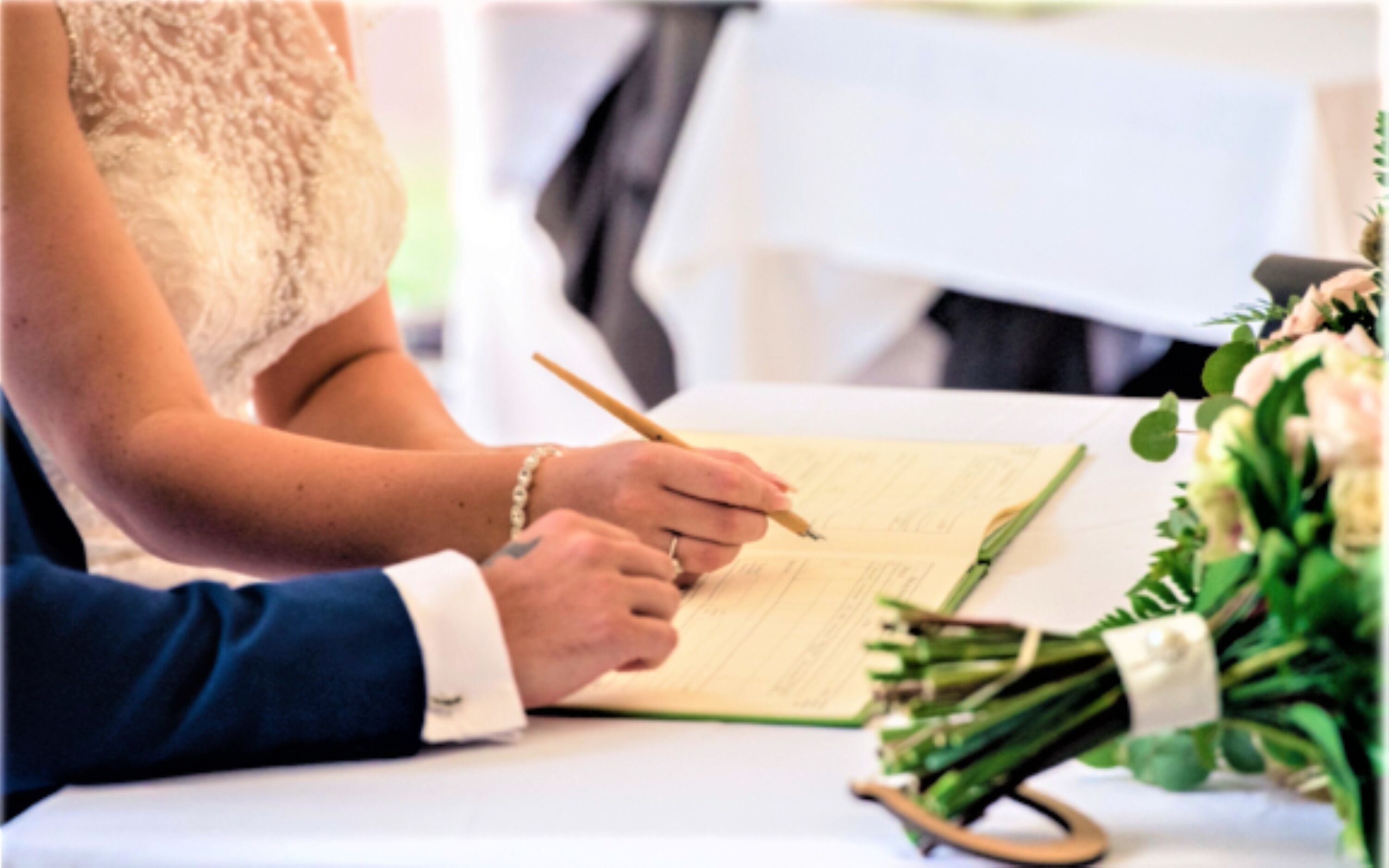 A person writing in a notebook with a pen, a bouquet of flowers, and a ring on their finger.