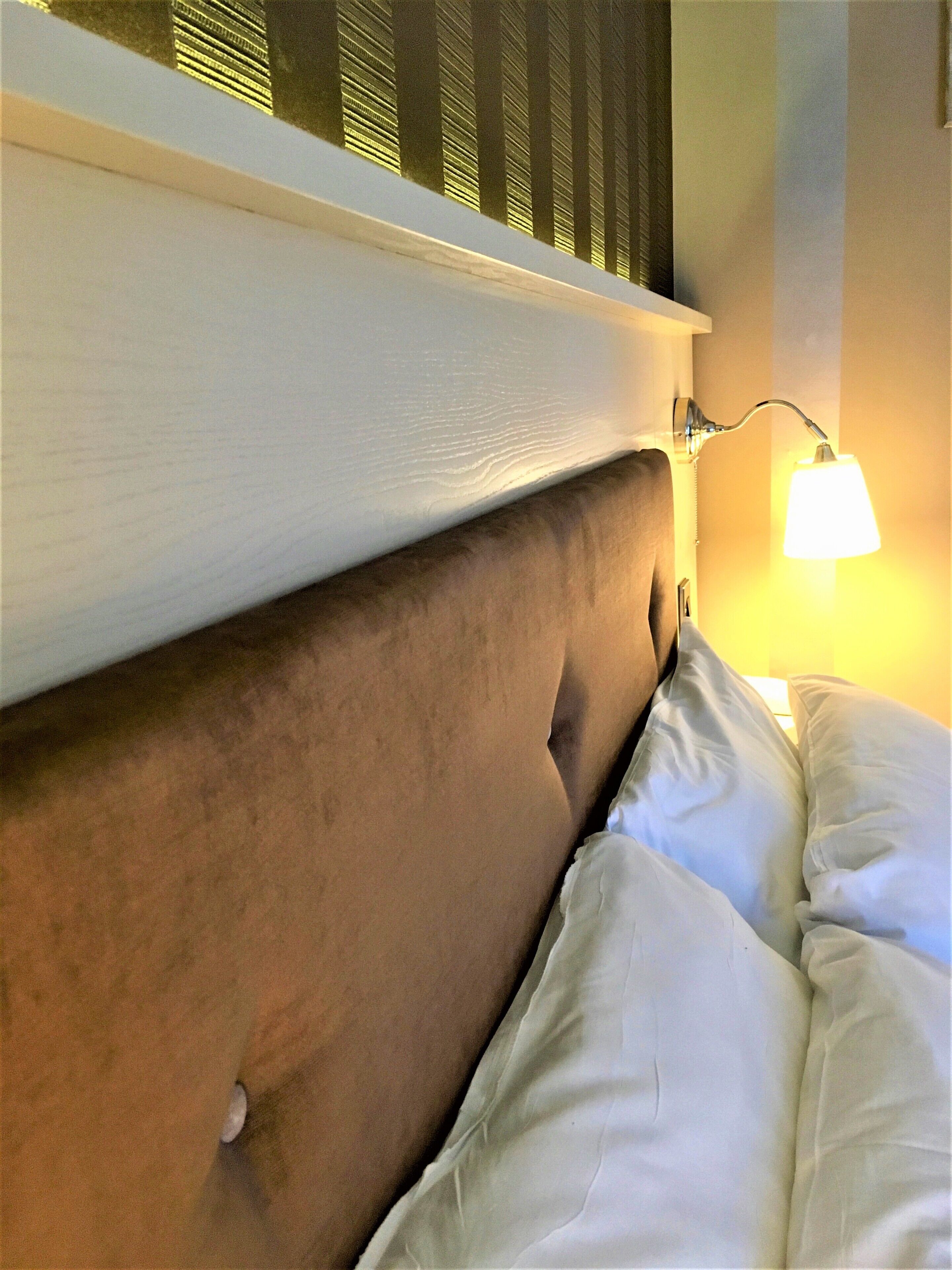 Una cama con cabecera, una lámpara de pared y un ventanal con persianas.