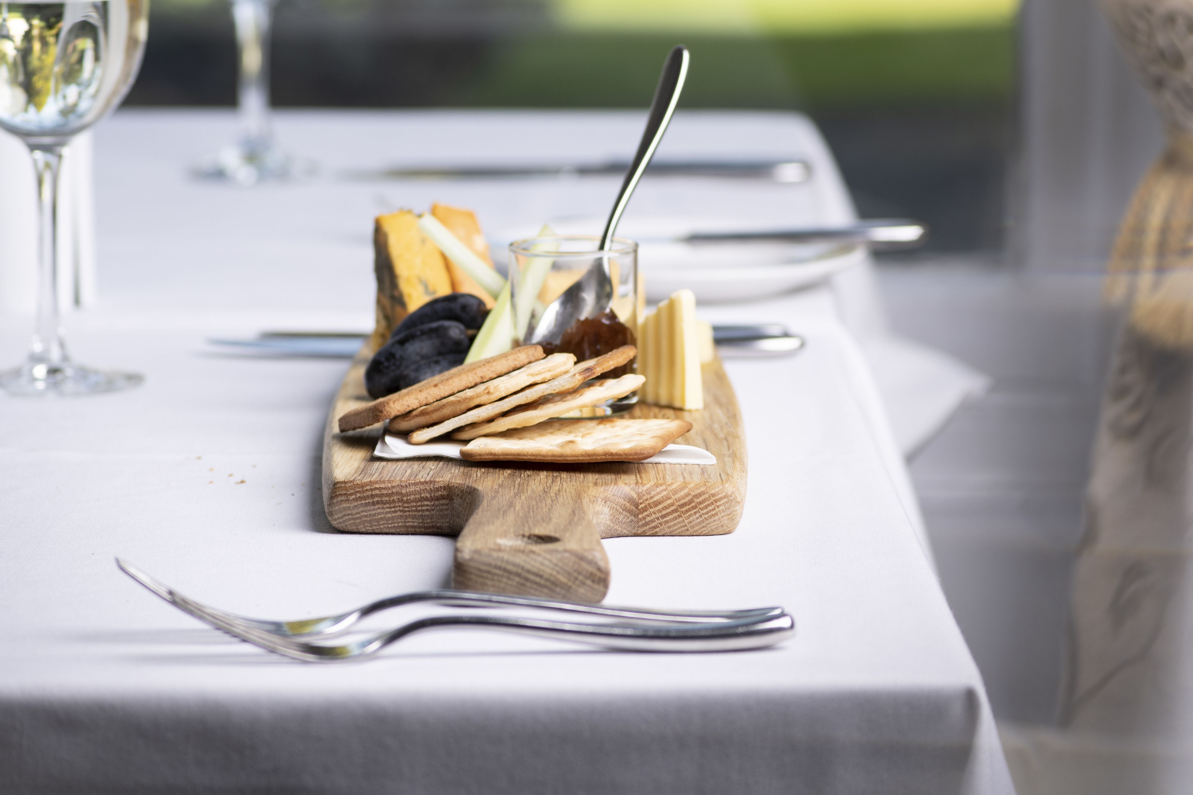A table set with a wooden board holding various snacks, a glass with a spoon, and cutlery.