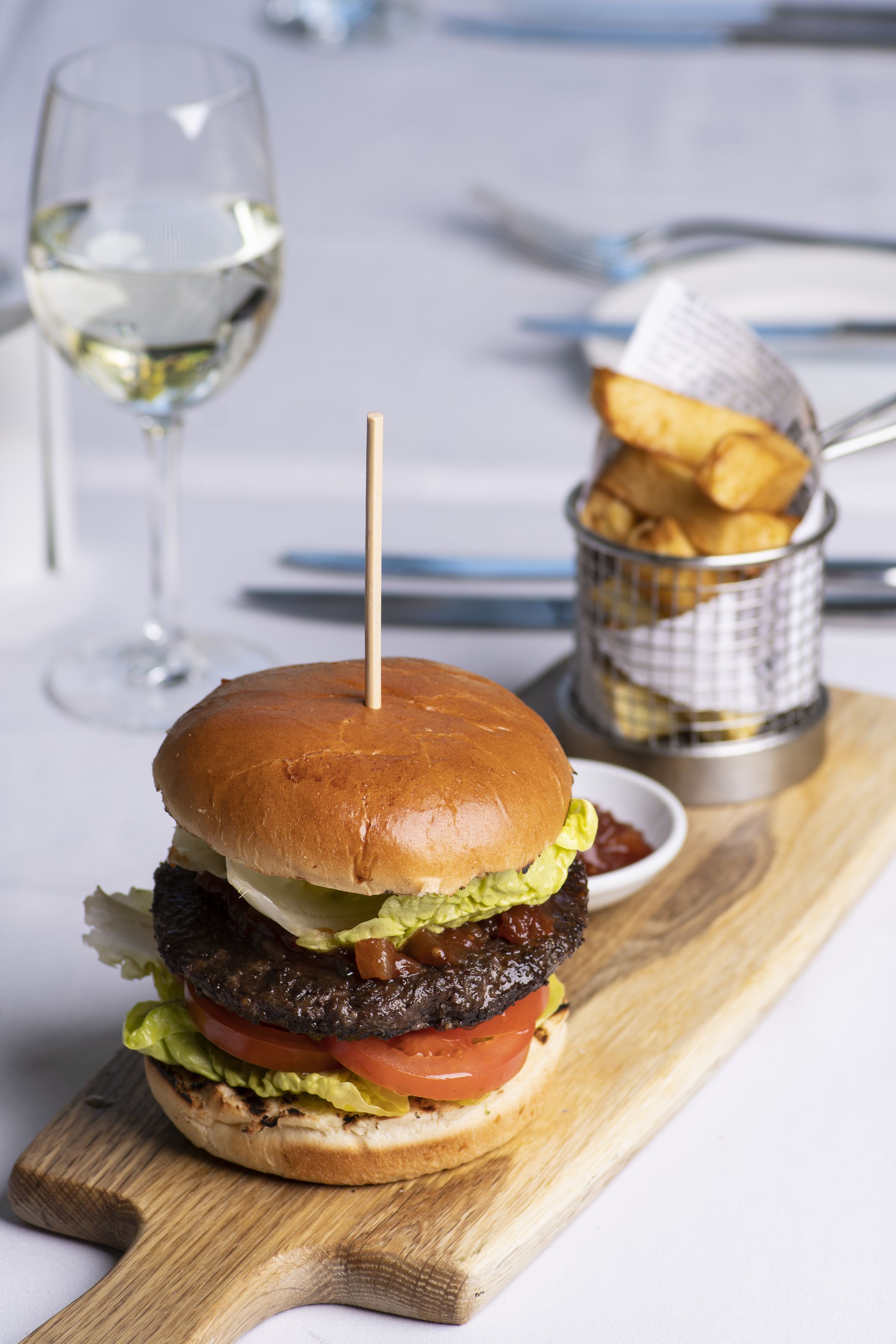 Un hamburger maintenu par un cure-dent, un verre de vin blanc et un panier de frites, le tout posé sur une planche en bois.
