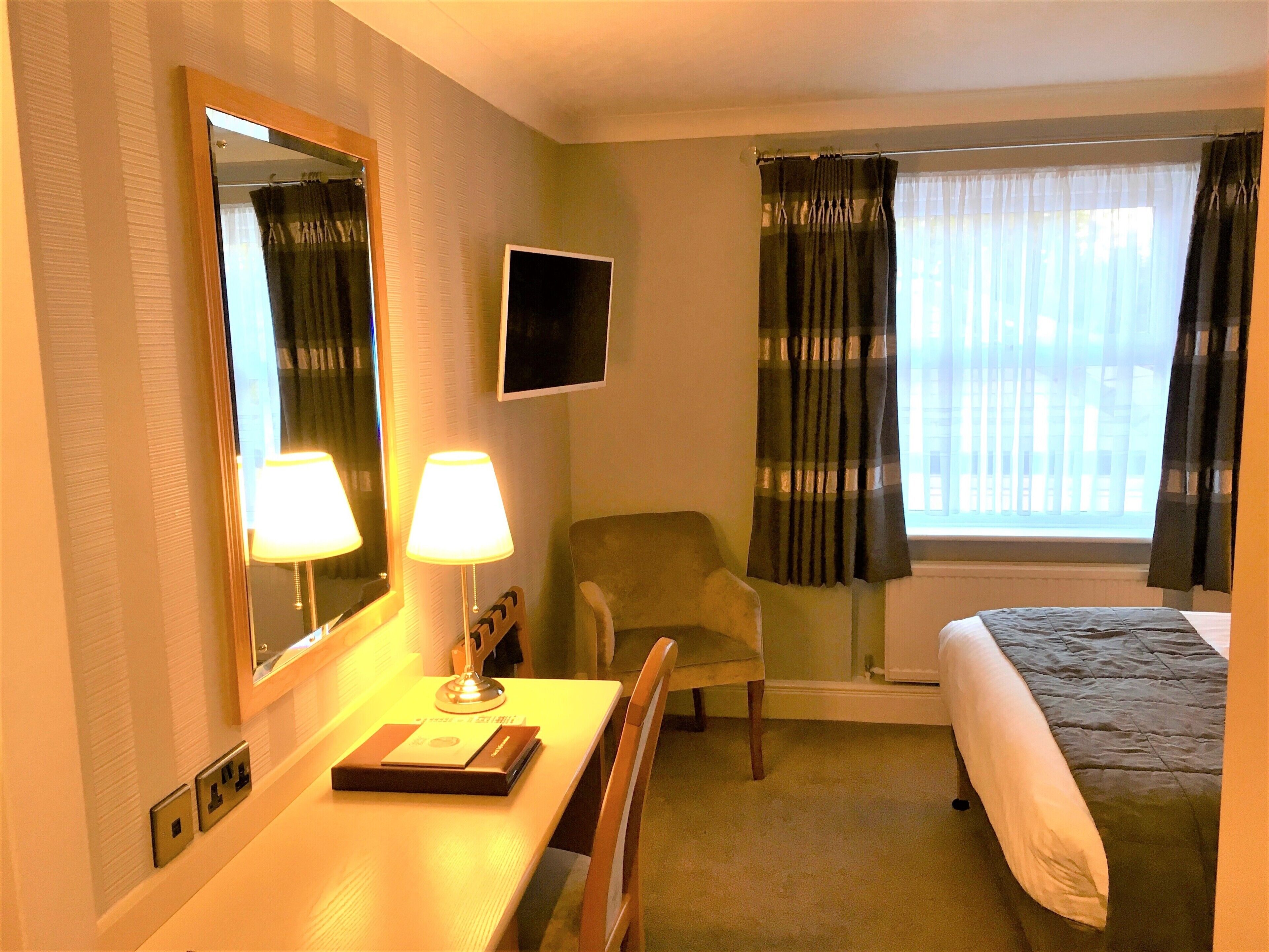 A hotel room with a bed, a desk with a lamp, a chair, a TV on the wall, and a window with curtains.