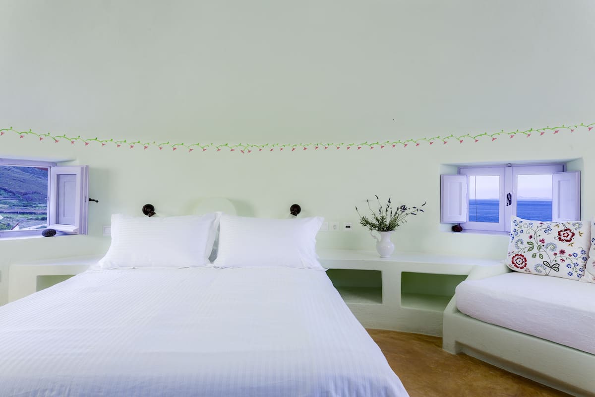 A bedroom with a large bed, two bedside tables, a small bedside table with a plant, and a window with a view.