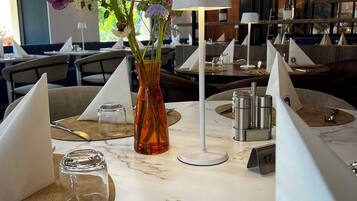 A restaurant setting with tables set for dining, featuring a vase with flowers, a lamp, and a menu board in the background.