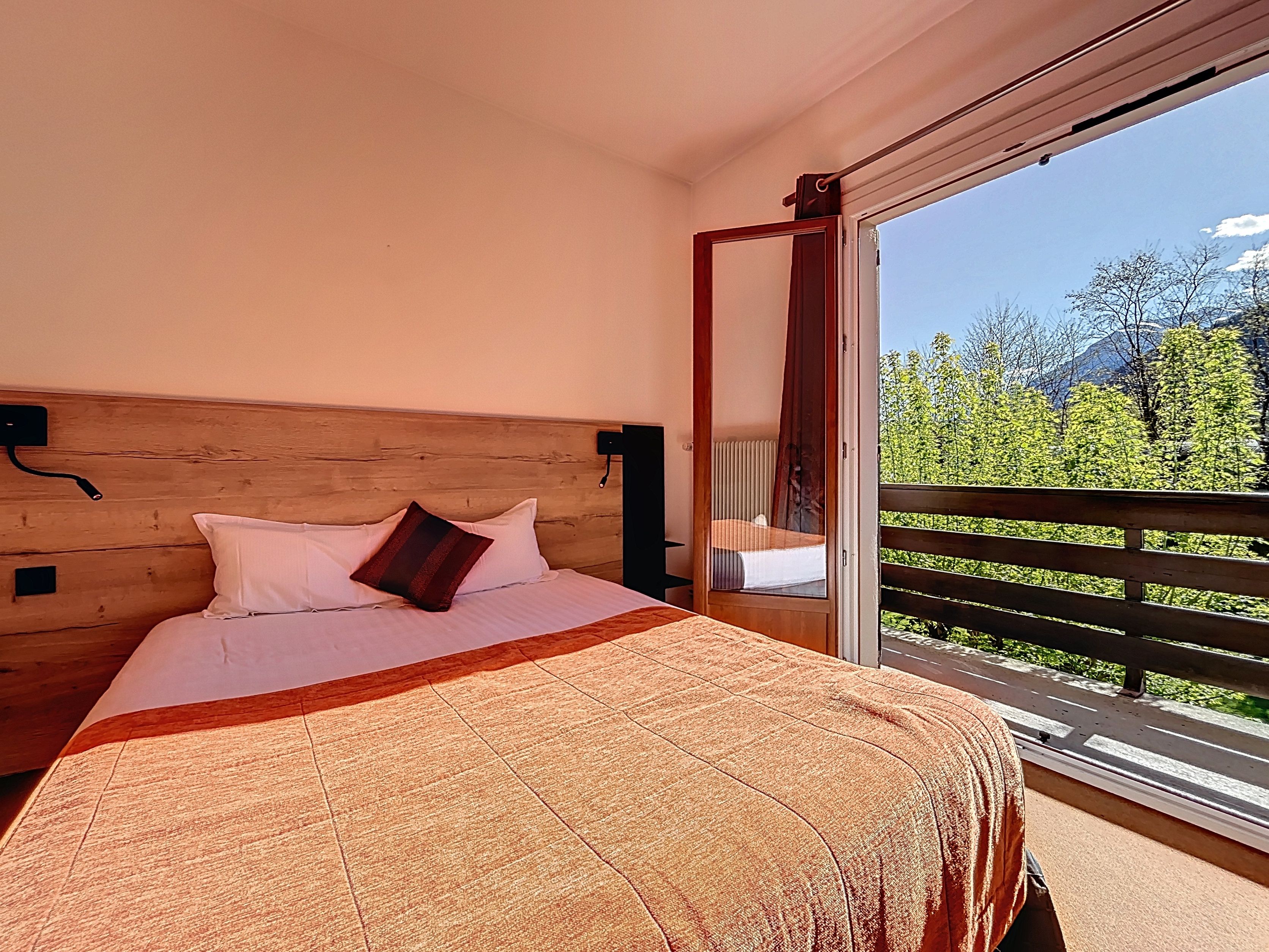 Un dormitorio con cabecera de madera, cama con ropa de cama blanca y una manta estampada, y un balcón con vistas a zonas verdes y montañas.